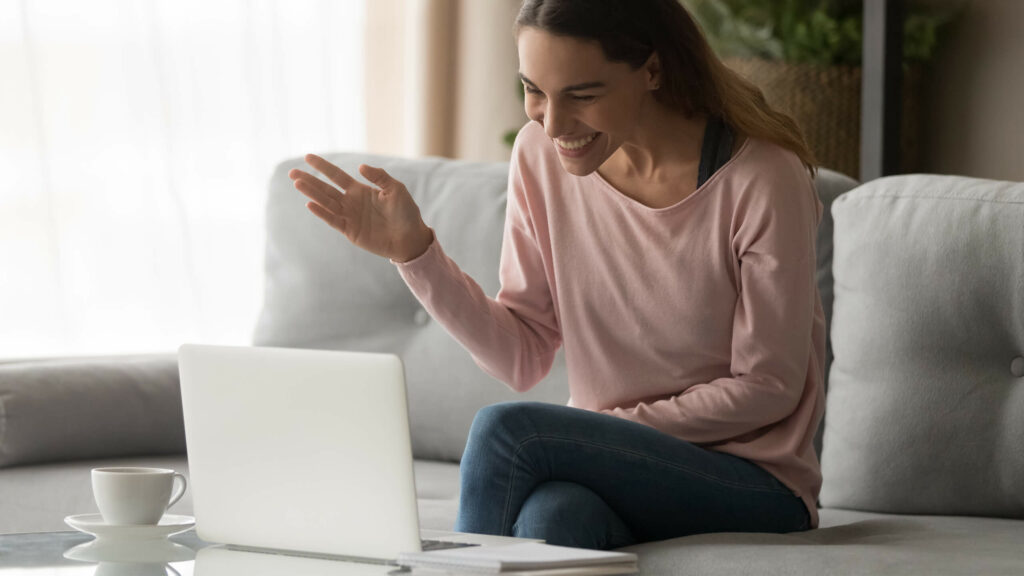 A woman smiling and waving at a laptop placed on the coffee table in front of her