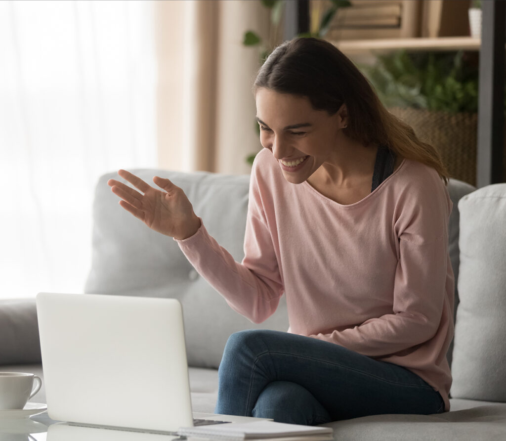 A woman smiling and waving at a laptop placed on the coffee table in front of her