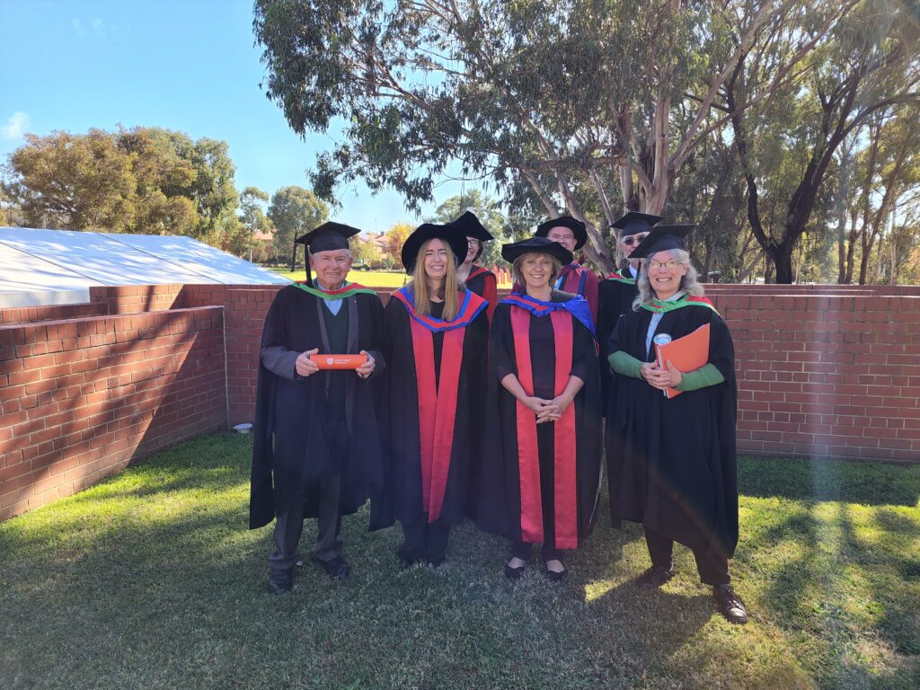 Seven men and women wearing academic gowns and smiling against a backdrop of eucalypts.